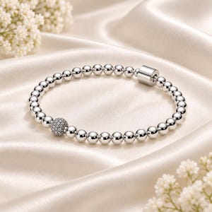 Puede incluir: Una pulsera de cuentas plateadas que presenta una serie de cuentas esféricas lisas y reflectantes, una cuenta texturizada incrustada con pequeñas piedras transparentes y un cierre cilíndrico, todo dispuesto en un diseño circular.