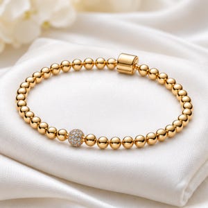 May include: A gold-toned beaded bracelet featuring a series of polished spherical beads and a single accent bead encrusted with small clear stones. The bracelet includes a cylindrical gold-toned clasp mechanism to secure the circular band.