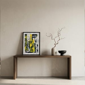 Puede incluir: Una pintura abstracta enmarcada de forma rectangular con trazos verticales de pintura amarilla, negra y blanca. La obra se exhibe sobre una consola de madera marrón oscuro junto a un jarrón de cerámica claro con una rama fina y desnuda.