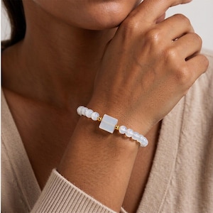 May include: A beaded bracelet featuring a series of smooth, translucent white spherical beads interspersed with small, polished gold-toned metallic spheres and a single central white square-shaped bead component.