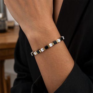 Può includere: Un bracciale caratterizzato da un motivo ripetitivo di perline quadrate nere, perline bianche con venature marmorizzate e piccoli distanziatori in metallo color oro. Le perline sono disposte in una sequenza lineare e strutturata.