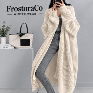 May include: An off-white, chunky knit open-front cardigan featuring long sleeves, ribbed cuffs, and two large front patch pockets. The garment is worn over a grey top and grey denim jeans. The text FrostoraCo WINTER WEAR is visible.