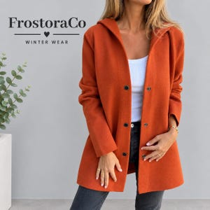 Puede incluir: Un abrigo con capucha de color naranja quemado que presenta un cierre frontal con botones oscuros. El texto FrostoraCo WINTER WEAR es visible en la esquina superior izquierda. El abrigo se lleva sobre una camiseta blanca y vaqueros gris oscuro.