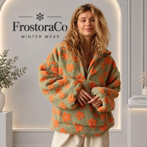 Puede incluir: Un suéter de forro polar verde salvia con un patrón floral naranja texturizado, cuello alto con cremallera parcial y mangas largas. La prenda se muestra frente a una pared con el texto FrostoraCo WINTER WEAR impreso en la parte superior.
