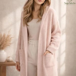 May include: A long, pale pink knit cardigan featuring a hood, long sleeves, and two front patch pockets. The garment is worn over a matching pale pink top and light-coloured trousers. The SageStitchs logo is visible in the upper right corner.