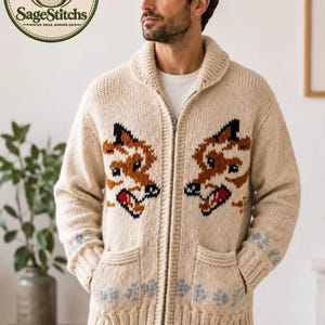 May include: A cream-colored knit zip-up cardigan featuring a shawl collar and two front pockets. The garment displays a brown and black fox head pattern on each side of the zipper, with light blue paw print accents near the ribbed cuffs and hem.