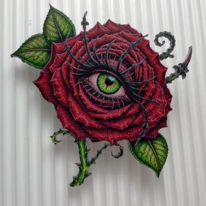 May include: A decorative wall art piece featuring a deep red rose with a realistic green eye at its center. The rose has long black eyelashes, green leaves with visible veins, and a thorny stem. The petals are detailed with small water droplets.
