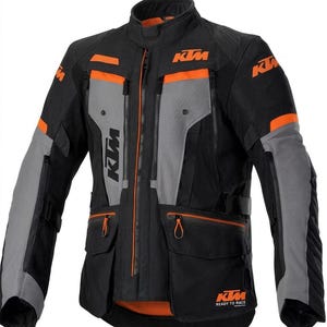 May include: A black, grey, and orange motorcycle jacket featuring the KTM logo on the chest, shoulders, and lower front. The jacket includes a high collar, multiple zippered pockets, adjustable straps on the sides, and the text READY TO RACE.