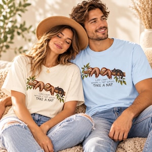 May include: A cream-coloured t-shirt and a light blue t-shirt, both featuring a graphic of two sloths and a black cat resting on a tree branch with the text: LIFE'S SHORT. NOTHING IS THAT SERIOUS. TAKE A NAP. The shirts have short sleeves and crew necks.