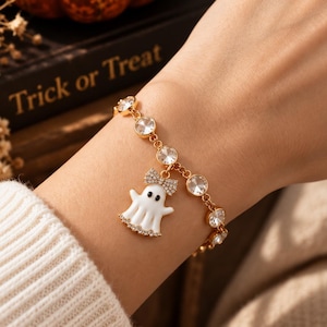 Könnte beinhalten: Ein goldfarbenes Kettenarmband mit runden, klaren Kristallen und einem zentralen weißen Geisteranhänger. Der Geisteranhänger ist mit einer kleinen, mit Kristallen besetzten Schleife am Kopf und einer Reihe winziger Kristalle am Saum verziert.