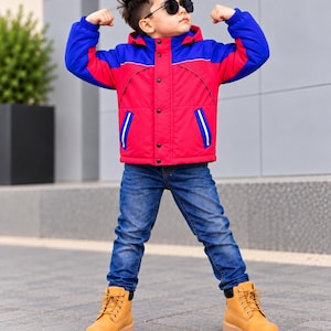 Könnte beinhalten: Eine Kapuzenjacke mit leuchtend rotem Rumpf sowie blauen Ärmeln und Schultern, kombiniert mit einer dunkelblauen Jeans und beigefarbenen Schnürstiefeln aus Leder. Die Jacke hat eine Knopfleiste, zwei Seitentaschen mit weißem Rand und eine schwarze Sonnenbrille.