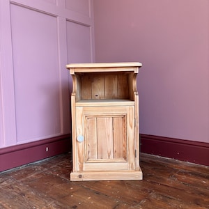 May include: A light brown wooden bedside table featuring a natural wood grain finish. The piece includes an open storage shelf at the top and a single cabinet door below with a small, round, light-coloured knob handle for opening the compartment.