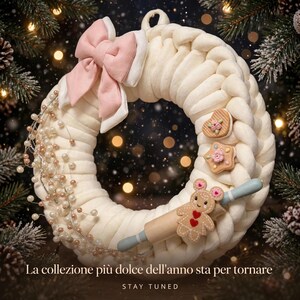 Puede incluir: Una corona de tela trenzada de color crema decorada con un gran lazo rosa pálido, un racimo de pequeñas cuentas blancas y oro rosa, y tres adornos: un corazón, una estrella y un hombre de jengibre sosteniendo un rodillo de madera.