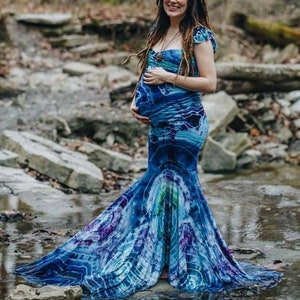 Puede incluir: Una persona embarazada con un vestido maxi azul y morado tie-dye con cola de sirena. El vestido tiene un escote de coraz&oacute;n y mangas cortas. La persona est&aacute; de pie en un arroyo con rocas y agua.