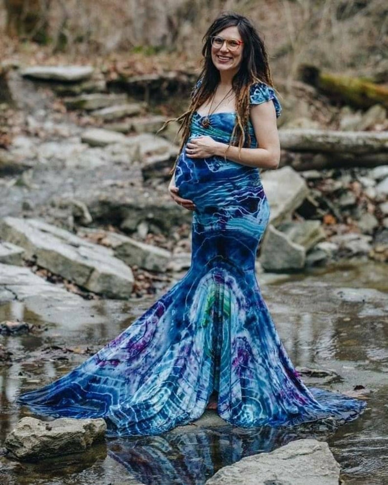 Puede incluir: Una persona embarazada con un vestido maxi azul y morado tie-dye con cola de sirena. El vestido tiene un escote de coraz&oacute;n y mangas cortas. La persona est&aacute; de pie en un arroyo con rocas y agua.