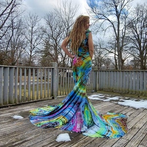 Puede incluir: Una mujer lleva un vestido largo y fluido de te&ntilde;ido anudado con un dise&ntilde;o de cola de sirena. El vestido es una mezcla de colores azules, verdes, rosas y amarillos. La mujer est&aacute; de pie en una plataforma de madera con nieve en el suelo.