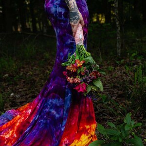 Puede incluir: Una mujer con un vestido de tie-dye colorido con una larga cola. Ella sostiene un ramo de flores en su mano.