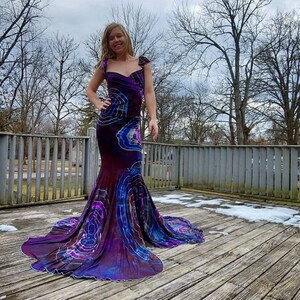 Puede incluir: Una mujer lleva un vestido largo de color morado y azul con estampado tie-dye y una falda de cola de sirena. El vestido tiene un escote de coraz&oacute;n y tirantes finos. La mujer est&aacute; de pie en una plataforma de madera con nieve en el suelo.