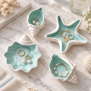 Puede incluir: Un conjunto de cuatro platos de cerámica para joyas con forma de conchas marinas y una estrella de mar, con exterior blanco e interior azul claro brillante. Los platos contienen varias piezas de joyería de oro y perlas, incluyendo anillos y pulseras.