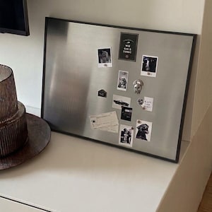 Può includere: Una bacheca magnetica rettangolare color argento con una cornice scura. La bacheca mostra varie piccole fotografie, appunti cartacei e un cartoncino rettangolare con il testo WIE GIN & TONIC, fissati da piccoli magneti circolari.