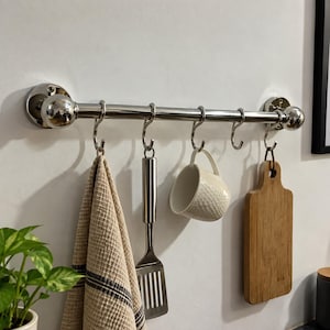 Puede incluir: Una barra de cocina de cromo pulido montada horizontalmente con cinco ganchos en S a juego. Colgados de los ganchos hay un paño de cocina beige texturizado, una espátula ranurada de acero inoxidable, una taza de cerámica blanca y una tabla.