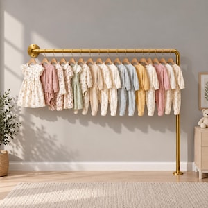 Puede incluir: Un perchero de metal dorado montado en la pared y el suelo, que exhibe una fila de prendas infantiles que incluyen vestidos, peleles y pijamas de manga larga con pies en varios tonos pastel, estampados florales y colores neutros.