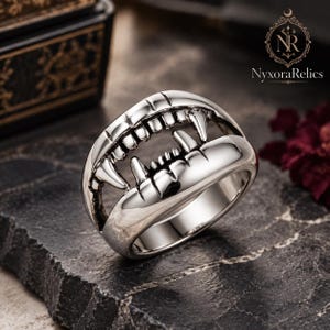 May include: A silver-toned ring featuring a sculpted design of an open mouth with prominent fangs and teeth. The metal surface is polished with dark recessed detailing to define the individual teeth and gum line, creating a realistic dental structure.