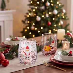Könnte beinhalten: Zwei klare doppelwandige Gläser mit einem weihnachtlichen Weihnachtsmann-Motiv und eingebetteten roten und grünen Glitzerpartikeln in den Glaswänden. Die Gläser haben eine abgerundete, stiellose Form für festliche Getränke.