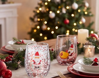 May include: Two clear double-walled glass tumblers featuring a festive Santa Claus graphic and embedded red and green glitter particles within the glass walls. The glasses have a rounded, stemless shape and are designed for holiday beverage service.