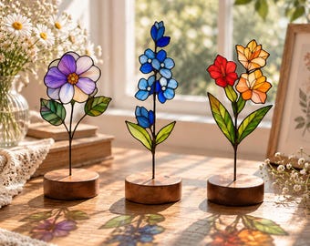 May include: Three stained glass flower sculptures on circular wooden bases. The left features a purple and white bloom, the center a tall blue stalk, and the right a cluster of red and orange blossoms, all casting colorful shadows on the surface.