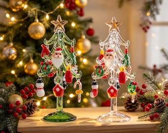 Op de afbeelding: Twee glazen kerstboomornamenten, één groen en één transparant, elk bekroond met een gouden ster. De bomen zijn versierd met miniatuur glazen bedeltjes, waaronder kerstmangezichten, sneeuwpoppen, zuurstokken en kransen die aan de takken hangen.