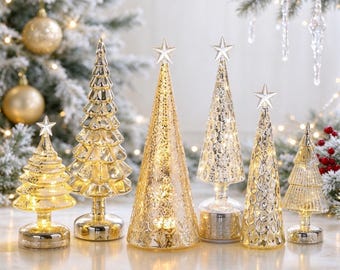 May include: A collection of six illuminated glass Christmas tree figurines in varying heights and textures. Each tree features a gold or silver metallic finish, a conical shape, and a small star topper, all glowing with internal warm LED lights.