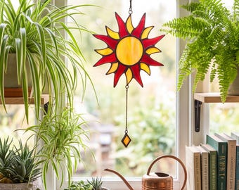 May include: A stained glass sun catcher featuring a central yellow circle surrounded by alternating red and yellow pointed rays. The piece hangs from a black metal hook and includes a thin chain with a small diamond-shaped glass pendant at the bottom.