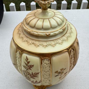 May include: A cream-colored ceramic jar with a matching lid, featuring gold-painted accents and intricate relief patterns. The body displays detailed brown botanical illustrations of ferns and flowers, separated by vertical gold-trimmed panels.