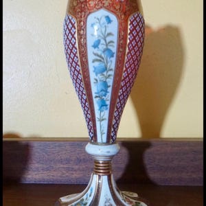 May include: A tall, ornate vase featuring a flared rim, a central panel decorated with blue bell-shaped flowers, and side panels with a red and white diamond lattice pattern. The base is white with gold accents and small floral motifs.