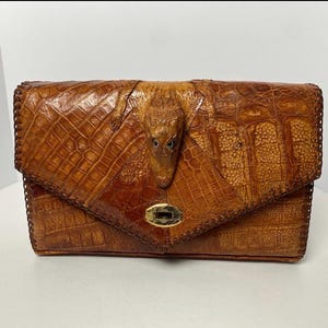 May include: A brown textured leather clutch bag featuring a prominent embossed alligator head on the front flap. The bag has a triangular envelope-style closure with a gold-tone metal turn-lock mechanism and decorative whip-stitched edges.