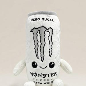 May include: A white plush toy shaped like a Monster Energy Ultra White drink can. The plush features a grey top, embroidered black Monster logo, and the text ZERO SUGAR and MONSTER ENERGY ULTRA WHITE. It has small arms, legs, and a cute face.