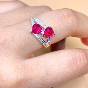 Puede incluir: Un anillo de tono plateado con dos piedras rojas vibrantes en forma de corazón colocadas una al lado de la otra. La banda está dividida en dos secciones, ambas incrustadas con pequeñas piedras claras y brillantes a lo largo del aro.