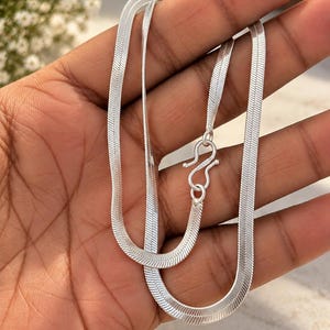 Puede incluir: Un collar de cadena de espiga en tono plateado con un diseño de eslabones planos y flexibles con un acabado metálico pulido. El collar se asegura con un mecanismo de cierre de gancho en S, mostrando una textura de superficie lisa y reflectante.