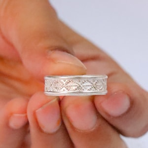 Può includere: Un anello a fascia in argento caratterizzato da un motivo ripetitivo a forma di diamante inciso lungo il centro. Il metallo presenta una finitura lucida con dettagli testurizzati all'interno di ogni rombo, creando un design geometrico uniforme.
