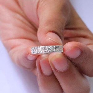Può includere: Un anello a fascia color argento caratterizzato da un motivo in rilievo ripetuto di piccoli fiori e foglie sulla superficie esterna. Il metallo presenta una finitura opaca e una larghezza costante, con dettagli floreali intricati lungo tutto il perimetro.