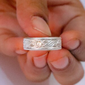May include: A silver-toned band ring featuring a continuous embossed pattern of small flowers and swirling leaves. The metal surface has a polished finish, and the design wraps entirely around the circumference of the circular ring band.