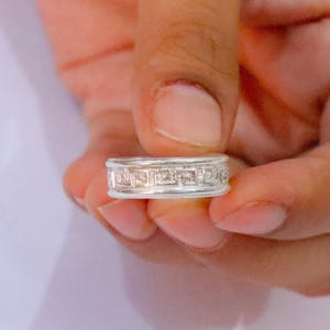 May include: A silver-toned band ring featuring a repeating geometric Greek key pattern engraved around the centre. The ring has a polished finish with raised edges on the top and bottom, creating a distinct border around the central design.