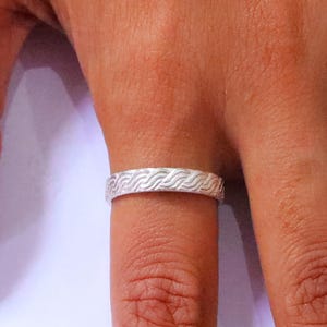 May include: A silver-coloured band ring featuring a continuous embossed pattern of repeating diagonal wave lines. The metal surface has a matte finish, and the ring is worn on a finger, showcasing the detailed texture of the engraved design.