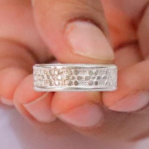May include: A silver-toned band ring featuring a textured surface with a repeating pattern of small, circular indentations across the entire outer circumference. The ring has a flat profile with smooth, raised edges along the top and bottom borders.