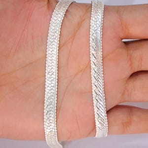 May include: A silver-toned flat chain necklace featuring a repeating woven or braided pattern across its entire length. The chain has a polished, reflective surface with scalloped edges along both sides, creating a textured appearance.