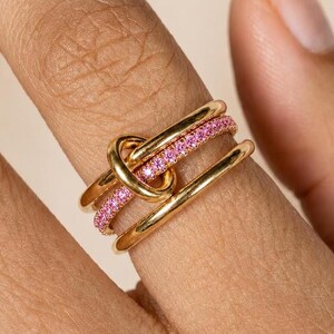 Könnte beinhalten: Ein goldener Ring bestehend aus einer polierten Schiene und einer zweiten Schiene, die mit kleinen rosa Steinen besetzt ist. Beide Ringe sind durch eine zentrale goldene Schlaufe verbunden und bilden ein geschichtetes Design mit glatter Oberfläche.