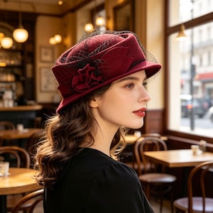 May include: A deep burgundy felt hat featuring a structured crown, a short brim, and a decorative fabric flower on the side. The hat is adorned with a fine black netting veil that drapes over the top and sides, creating a textured appearance.