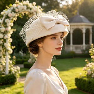 May include: A cream-coloured felt pillbox hat featuring a structured bow on the front and a delicate, fine-mesh netting veil that drapes over the top. The hat is crafted from a smooth, solid material and includes a small pearl accent on the side.