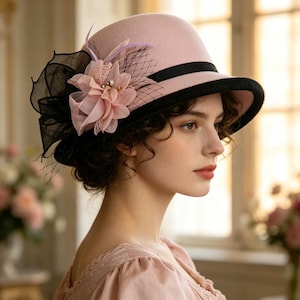 Può includere: Un cappello in feltro rosa chiaro con tesa nera e nastro nero. Il lato è decorato con un grande fiore in tessuto rosa a strati, rete nera e sottili piume nere, creando un accessorio strutturato e ricco di dettagli.